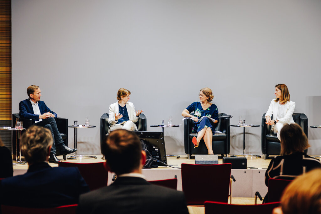 Foto einer Podiumsdiskussion. Vier Personen sitzen in dunklen Sesseln mit Beistelltisch und unterhalten sich. Die Personen schauen sich an. Die Personen tragen förmliche Kleidung. Im Vordergrund sind die Hinterköpfe von Personen aus dem Publikum zu sehen.