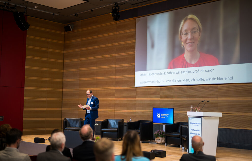 Foto von der CDR-Konferenz 2023. Der Moderator steht auf einer Bühne. Im Hintergrund ist eine Person online zugeschaltet. Vor der Bühne sitzt ein Publikum.